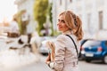 Enchanting blonde girl in trendy beige jacket having fun outside walking by cars. Outdoor portrait of enthusiastic
