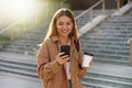 Enchanting blonde young woman waiting for phone message while carrying coffee cup on the street. Stylish girl in spring clothing