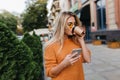 Enchanting blonde young woman waiting for phone message while drinking coffee on the street. Stylish girl in yellow