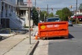 The end of an orange dumpster seen on a street in front of a truck at a construction site on a sunny day