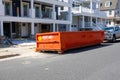 The end of an orange dumpster seen on a street in front of a truck at a construction site on a sunny day