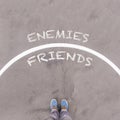 Enemies vs Friends text on asphalt ground, feet and shoes on flo