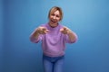 energetic cute blond young woman in purple sweater pointing with hand at camera on blue background with copy space