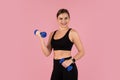Energetic smiling woman exercising with blue dumbbells against pink studio background