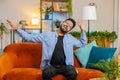 Energetic young Indian man singing dancing with hands sitting on couch room enjoying listening music Royalty Free Stock Photo