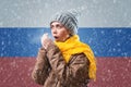Energy crisis. Russian woman in hat, coat and scarf is warming herself. In the background is the flag of Russia and a Royalty Free Stock Photo