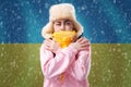 Energy crisis. Ukrainian woman in hat, coat and scarf is warming herself. In the background is the flag of Ukraine and a Royalty Free Stock Photo