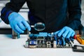 Engineer in blue gloves inspecting a computer motherboard with a magnifying glass in a tech lab. Concept of electronics repair, Royalty Free Stock Photo