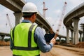 Engineer in high-visibility vest and hard hat using tablet at highway construction site with cranes bridge Royalty Free Stock Photo