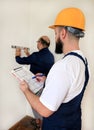 Engineer with his colleague, construction worker is measuring wall using spirit level tool, checking in apartment blueprint.