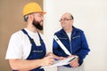 Engineer with his colleague, construction worker is measuring wall using spirit level tool, checking in apartment blueprint.