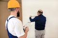 Engineer with his colleague, construction worker is measuring wall using spirit level tool, checking in apartment blueprint.