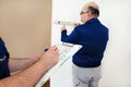 Engineer with his colleague, construction worker is measuring wall using spirit level tool, checking in apartment blueprint.