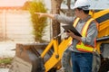 Engineering wearing a white safety helmet standing in front of the backhoe Looking at home construction work And use the tablet to Royalty Free Stock Photo