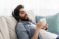 Enjoy morning at home. Relaxed arab millennial man in wireless headphones, reclining on sofa and drinking coffee
