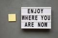 `Enjoy where you are now` words on lightbox over gray background, top view. Flat lay, from above