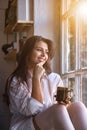 Portrait cute young woman in white shirt drinking tea and looking through window while sitting at windowsill indoors Royalty Free Stock Photo