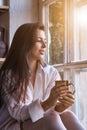 Portrait cute young woman in white shirt drinking tea and looking through window while sitting at windowsill indoors Royalty Free Stock Photo