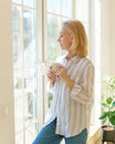 Beautiful senior woman with cup of coffee looking in window while enjoying new day at home Royalty Free Stock Photo