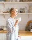 Beautiful senior woman with cup of coffee or rure water looking in window while enjoying new day at home, vertical Royalty Free Stock Photo