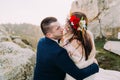 Enloved groom in stylish blue suit holding romantic white dressed bride with cute head wreath on majestic mountain rocky