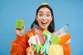 Enthusiastic asian girl showing cleaning sponge, holding empty plastic bottles, trash for recycling, sorting her