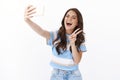 Enthusiastic happy woman posing near white background raise hand hold smartphone, taking positive cute selfie wink