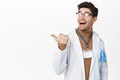 Enthusiastic male physician, happy doctor in glasses pointing and looking left, behind his shoulder, showing clinic