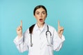 Enthusiastic medical worker, young woman doctor in white coat, stethoscope, showing advertisement, pointing fingers up