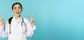 Enthusiastic medical worker, young woman doctor in white coat, stethoscope, showing advertisement, pointing fingers up