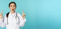 Enthusiastic medical worker, young woman doctor in white coat, stethoscope, showing advertisement, pointing fingers up