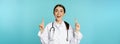 Enthusiastic medical worker, young woman doctor in white coat, stethoscope, showing advertisement, pointing fingers up