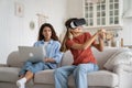 Enthusiastic positive teenage girl with VR glasses with virtual baseball bat sits on couch near mom Royalty Free Stock Photo