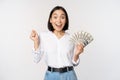 Enthusiastic young asian woman looking excited at camera, holding money dollars in hand, standing over white background Royalty Free Stock Photo