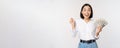 Enthusiastic young asian woman looking excited at camera, holding money dollars in hand, standing over white background Royalty Free Stock Photo