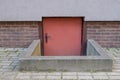 Entrance to basement with red door and metal handle fenced with border in lower part of gray decorative panel facade of building