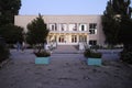 The entrance to the building is illuminated by bulbs under a visor with cracked asphalt near the entrance and flowers in