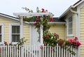 Entrance to cheery yellow wood house with white picket fence and a gate with an arbor with wild roses growing up and over it Royalty Free Stock Photo