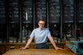 Entrepreneur wearing apron and glasses standing near counter of his cafe with big variety of coffee.