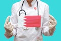 Epidemic in Bahrain. Young woman doctor in a medical coat and gloves holds a medical mask with the print of the flag of Bahrain on