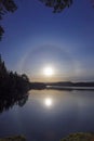 Ethereal lake vista with solar halo in blue sky and the water reflection of sunbeam, forest and mountains in Scandinavia. The circ Royalty Free Stock Photo