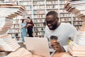 Ethnic african american guy surrounded by books in library. Student is using laptop and drinking coffee. Royalty Free Stock Photo