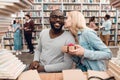 Ethnic african american guy and white girl surrounded by books in library. Students are giving gift. Royalty Free Stock Photo