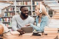 Ethnic african american guy and white girl surrounded by books in library. Students are using tablet. Royalty Free Stock Photo