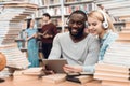 Ethnic african american guy and white girl surrounded by books in library. Students are using tablet. Royalty Free Stock Photo