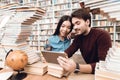 Ethnic asian girl and white guy surrounded by books in library. Students are using tablet. Royalty Free Stock Photo