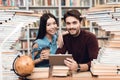 Ethnic asian girl and white guy surrounded by books in library. Students are using tablet. Royalty Free Stock Photo