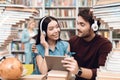 Ethnic asian girl and white guy surrounded by books in library. Students are using tablet with headphones. Royalty Free Stock Photo