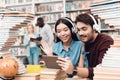 Ethnic asian girl and white guy surrounded by books in library. Students are using tablet with headphones. Royalty Free Stock Photo