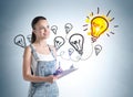 European attractive young woman in denim overalls holding planner and pondering near wall with colorful business idea lightbulb